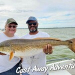 Large redfish caught during a Cast Away Adventures fishing trip with two smiling men on a boat by the water under a partly cloudy sky.